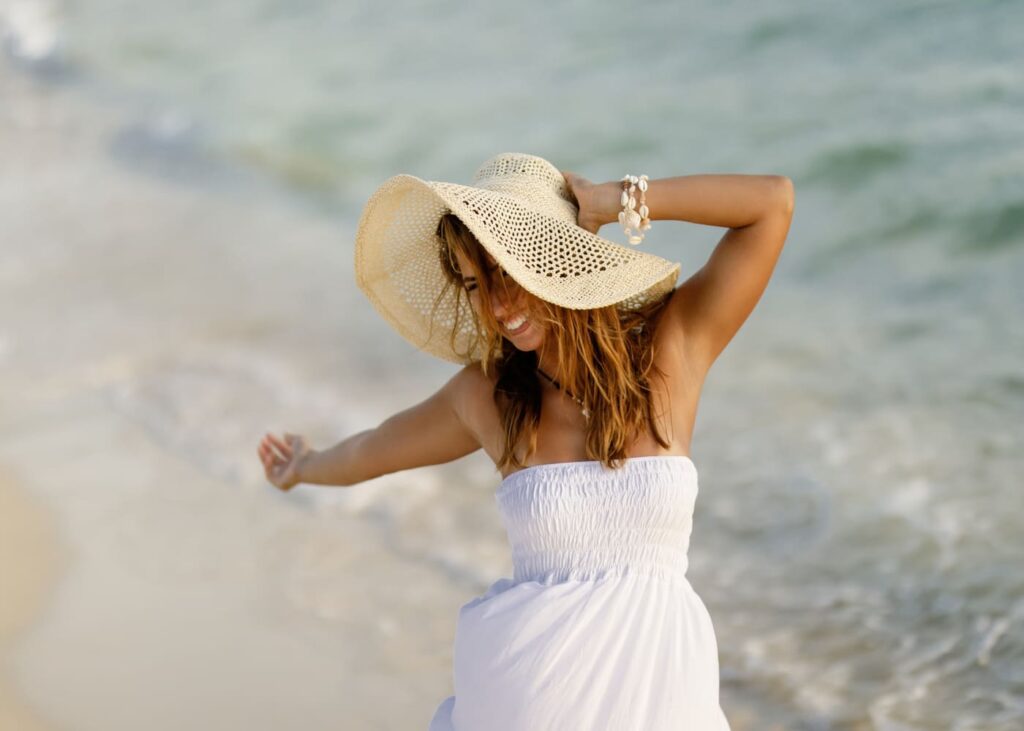 mujer-rubia-con-un-sombrero-de-paja-y-un-vestido-blanco-que-sonrie-frente-al-mar