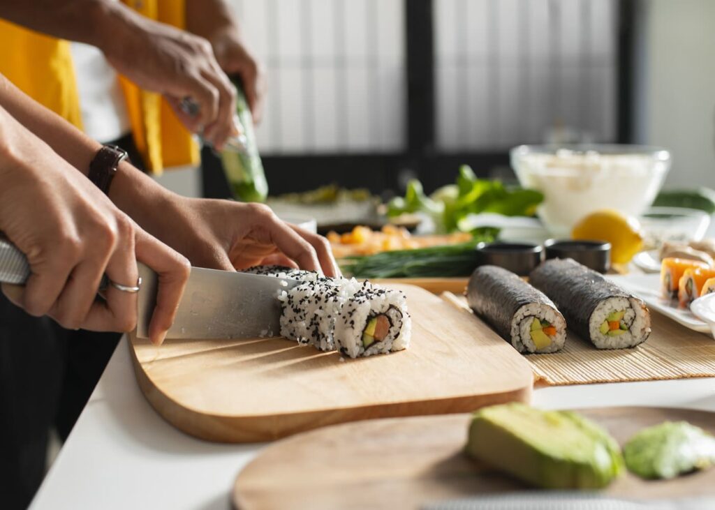 primer-plano-de-dos-manos-cortando-sushi-sobre-una-mesa-llena-de-ingredientes