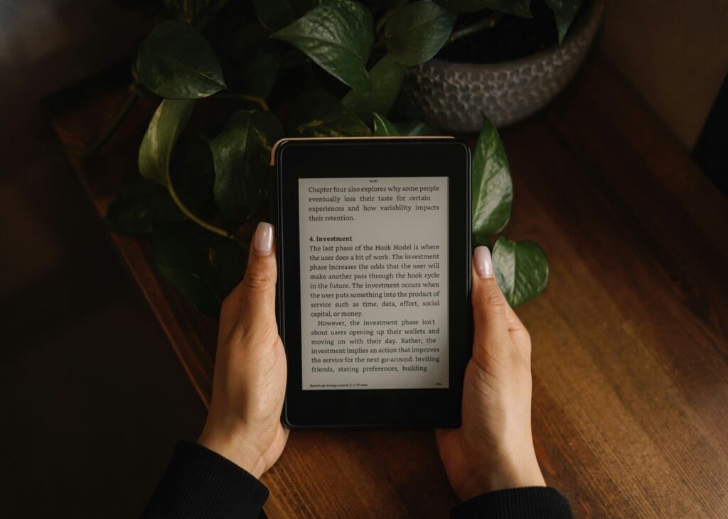 primer-plano-de-manos-de-mujer-que-sostiene-un-libro-electronico-con-mesa-de-madera-y-planta-al-fondo