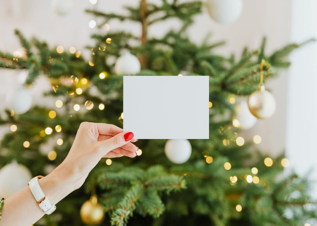 primer-plano-de-una-mano-de-mujer-con-esmalte-rojo-que-sostiene-una-tarjeta-blanca-frente-a-un-árbol-de-navidad