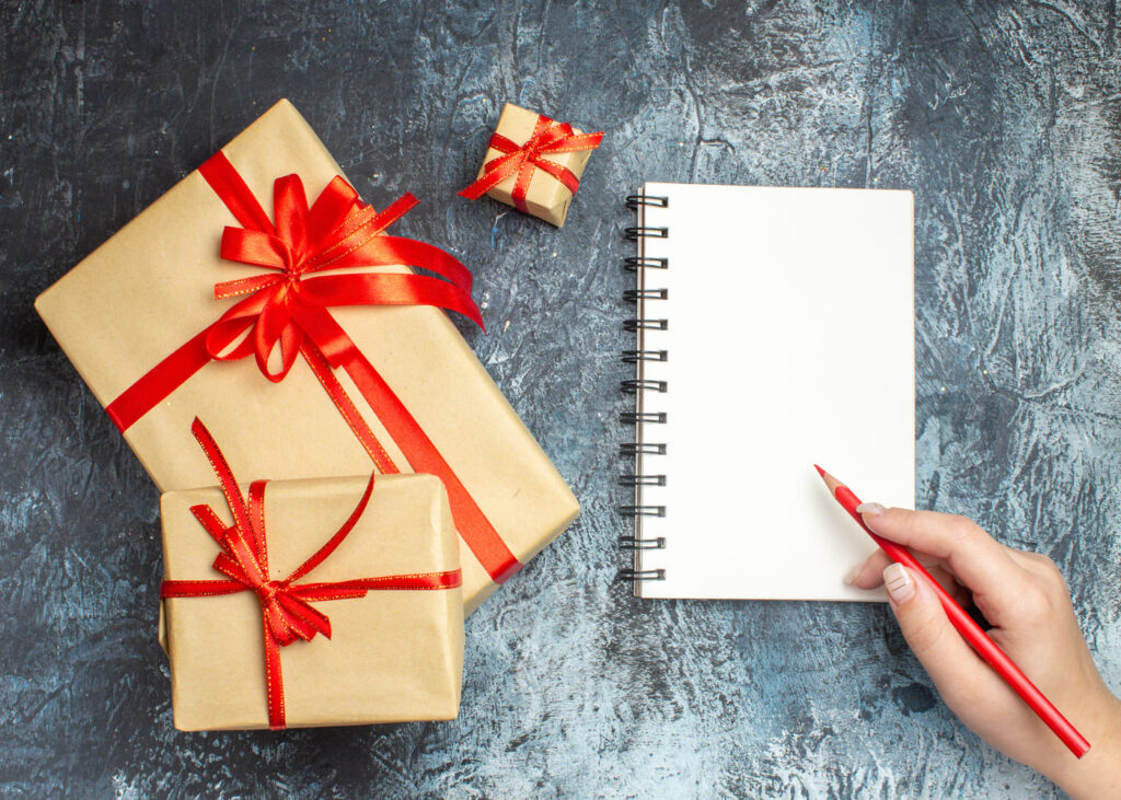 vista-desde-arriba-de-una-mano-que-escribe-en-un-cuaderno-y-regalos-al-lado-sobre-una-mesa-gris